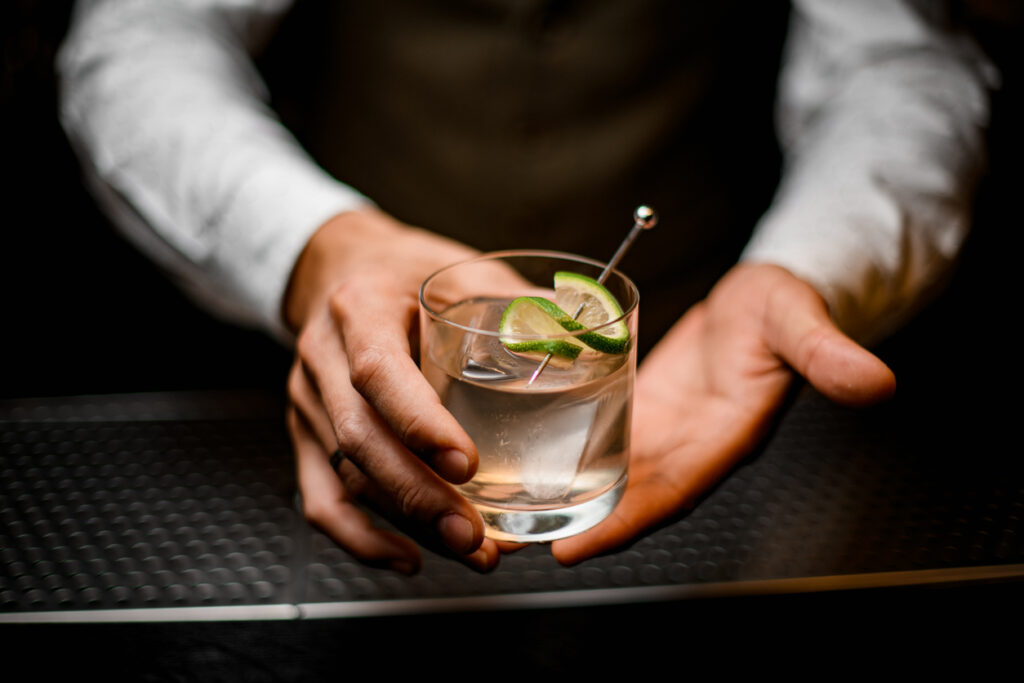 Cose-up on hands of man bartender holding glass with cold drink with ice cube decorated with lime. The kind that inspires many Drinks Buzzwords